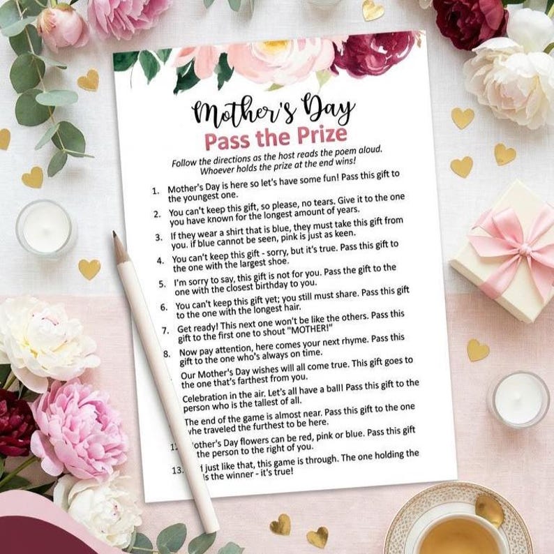 May include: A printed Mother's Day game sheet titled "Mother's Day Pass the Prize" with instructions and prompts. The sheet is surrounded by floral decorations and a pencil. A gift box and a cup of tea are also visible.