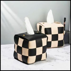 Puede incluir: Dos cubiertas de caja de pañuelos cuadradas con un diseño de tablero de ajedrez tejido. Una es negra y crema, la otra crema y negra. Cada una tiene un cierre de solapa y un pañuelo blanco que sobresale de la parte superior.