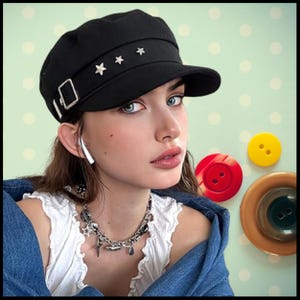 Puede incluir: Una gorra de newsboy negra con una hebilla plateada y detalles de estrellas. La gorra tiene una visera ligeramente curvada y la lleva puesta una persona con el pelo oscuro.