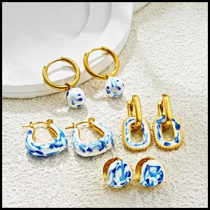 Op de afbeelding: Een verzameling goudkleurige oorbellen met wit en blauw gemarmerde emaille ontwerpen. De oorbellen omvatten oorringen, vierkante oorringen en oorknopjes. De sieraden worden gepresenteerd op een wit oppervlak.