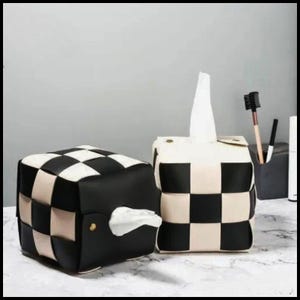 May include: Two tissue box covers with a woven black and cream checkerboard design. One is horizontal, the other vertical with tissues protruding. The boxes sit on a marble-effect surface.