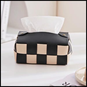 May include: A black and beige woven tissue box. The rectangular box has a black top flap with a gold-coloured button. White tissues protrude from the opening. The box is on a white surface, suitable for a desk or bedside table.