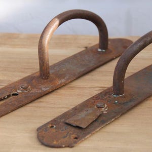 May include: Two antique, rusty metal door handles with a curved handle design. The handles are mounted on rectangular plates with keyhole and latch details. The metal has a weathered, aged appearance, suggesting vintage hardware.