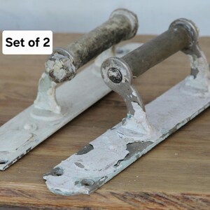 Puede incluir: Un juego de 2 manijas de puerta de metal con un acabado de pintura blanca desgastada y desconchada sobre una base oscura. Cada manija presenta una placa rectangular larga con orificios para tornillos y un agarre cilíndrico. Texto Set of 2.