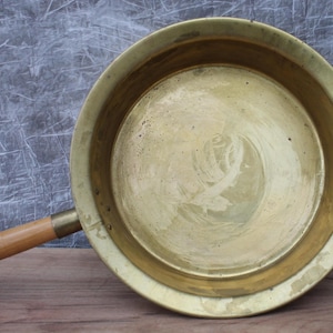 Op de afbeelding: Een koperen pan met een houten handvat. De pan is rond en heeft een glad, glanzend oppervlak. Het handvat is aan de zijkant van de pan bevestigd.