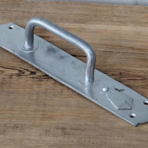 May include: A silver-colored door handle with a curved handle and a rectangular backplate. The backplate has decorative edges and a diamond-shaped design. The handle is mounted on a wooden surface.