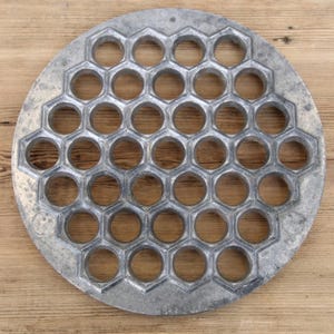 May include: A circular silver-toned metal pelmeni maker featuring a repeating honeycomb grid pattern with numerous evenly spaced circular cutouts throughout the surface, designed for shaping multiple dumplings at once.