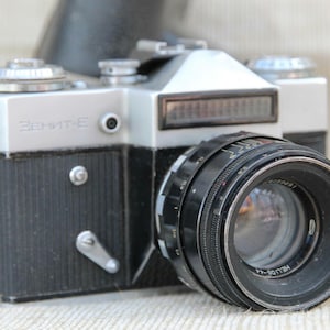 Può includere: Una fotocamera vintage argento e nera con un obiettivo prominente. Il corpo macchina presenta la scritta "Zenit-E". L'obiettivo è nero con segni bianchi. La fotocamera ha un flash e vari quadranti e leve.