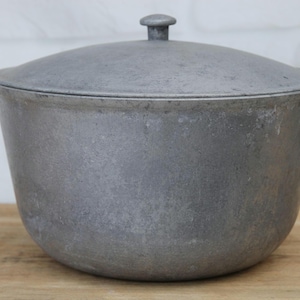 Könnte beinhalten: Ein grauer Kochtopf aus Gusseisen mit Deckel. Der Topf hat einen abgerundeten Körper, zwei Griffe und einen Deckel mit einem kleinen Knopf. Er steht auf einer Holzoberfläche.