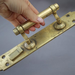 May include: Antique brass door handle with a long rectangular backplate. The handle is a cylindrical bar with curved supports. The brass has a warm, aged patina. The backplate has three screw holes and a rectangular lock mechanism.