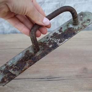 Może przedstawiać: Antyczna metalowa klamka do drzwi z zakrzywionym, zardzewiałym żelaznym uchwytem i prostokątną płytą tylną. Płyta tylna ma zniszczony, postarzany wygląd z zieloną i brązową oksydacją. Klamka jest trzymana przez osobę.