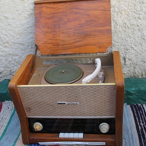 Puede incluir: Un tocadiscos vintage de madera con un gabinete de madera marrón y un panel frontal beige. El tocadiscos tiene un plato giratorio con un disco verde en él y una radio con una cara negra y perillas plateadas. El tocadiscos tiene el texto "Radio  Zira" en la parte delantera.