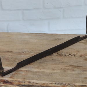 Pode incluir: Uma plaina de puxar vintage com uma lâmina de metal longa, reta e escura com um gume afiado. A ferramenta possui dois cabos cilíndricos de madeira fixados em cada extremidade, posicionados perpendicularmente à lâmina, apresentando sinais de desgaste.