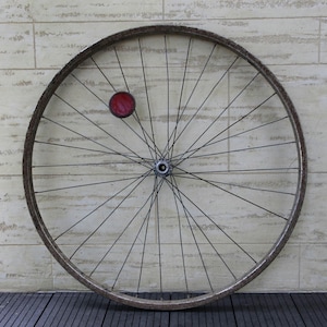 May include: A vintage bicycle wheel featuring a weathered, dark metal rim and numerous thin wire spokes radiating from a central hub. A single circular red reflector is attached to the spokes in the upper left quadrant of the wheel structure.