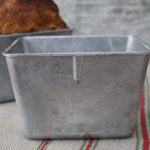 Puede incluir: Dos moldes para pan de metal plateado. El molde en primer plano está vacío y el molde en el fondo tiene una hogaza de pan dentro.