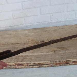 Pode incluir: Uma ferramenta de metal longa, estreita e enferrujada, com uma alça plana e retangular numa extremidade e uma barra de metal perpendicular e angular na extremidade oposta. A superfície do objeto de ferro apresenta oxidação acentuada.