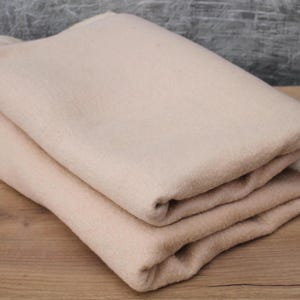 May include: A folded beige blanket made of soft, plush fabric. The blanket is folded in half and then folded again, creating a rectangular shape.