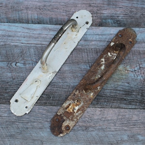 Puede incluir: Dos manillas de puerta antiguas, una blanca y otra marrón oxidada, con herrajes metálicos. Ambas manillas tienen un diseño simple y curvo.