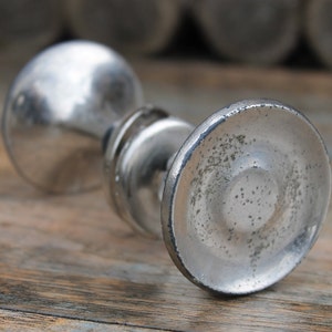Könnte beinhalten: Ein silberner Türknauf mit einem runden, leicht strukturierten Sockel und einem zylindrischen Kopf. Der Knauf hat ein abgenutztes, gealtertes Aussehen.