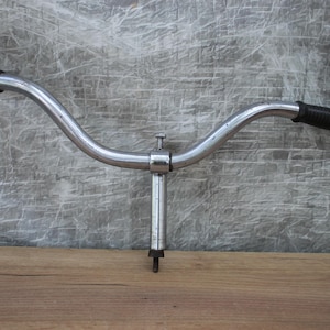 May include: A vintage bicycle handlebar with a curved design, made of silver-colored metal. The handlebar features black rubber grips on each end. The stem is vertical with a bolt for attachment. The background is a textured gray surface.