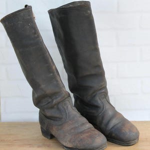 Puede incluir: Un par de botas altas de cuero marrón oscuro con punta redondeada, tacón plano y una caña estructurada que llega a media pantorrilla. El cuero muestra signos de desgaste con un acabado mate envejecido y costuras visibles en el empeine.