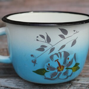 Op de afbeelding: Een blauwe emaille mok met een witte rand en een zwart bloemenontwerp. De mok heeft een witte handgreep en een blauwe kleurverloop.