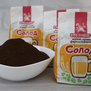 May include: A white bowl filled with dark brown malt powder is in front of three bags of malt. The bags have a red and white top with the text "СТО ПУДІВ" and a beer mug illustration. The bags also have the text "Солод".