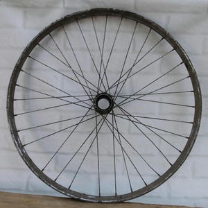 May include: A circular metal bicycle wheel featuring a dark, weathered rim and numerous thin, radiating spokes connected to a central hub. The metal surface shows signs of age, wear, and oxidation, with a visible dark finish throughout the structure.