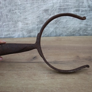 May include: An antique, rusty metal tool with a curved, open design. The tool has a handle held by a hand and a curved top. The tool is set against a wooden surface and a gray background.