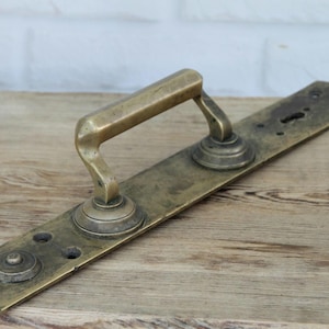 Op de afbeelding: Een vintage koperen deurklinkset met een lange rechthoekige achterplaat, een centrale hendel, een sleutelgat aan het ene uiteinde en een ronde knop aan het andere uiteinde. Het metaal heeft een verweerde patina met zichtbare schroefgaten.