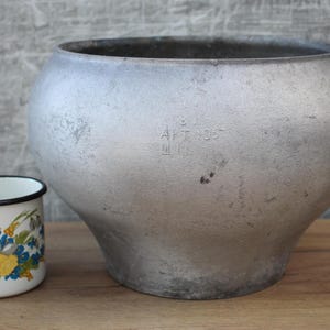 May include: A large, silver, round pot with a black rim and a stamped inscription that reads "AFT 1955". A small, white enamel mug with a floral pattern is in the foreground.