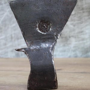 Pode incluir: Uma cabeça de machado de metal escuro e desgastado, com uma lâmina triangular e um cabo curvo. A cabeça do machado apresenta um emblema circular decorativo perto do topo. A ferramenta é exibida em uma superfície de madeira.