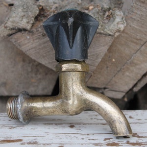 Può includere: Un rubinetto in ottone con una maniglia in plastica nera. Il rubinetto è su una superficie di legno bianco.
