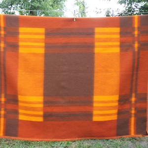 Può includere: Una coperta rettangolare con un motivo geometrico nei toni dell'arancione, marrone e giallo è appesa a un filo per stendere. La coperta ha un quadrato centrale marrone, circondato da strisce arancioni e gialle. La coperta è fatta di un materiale spesso.