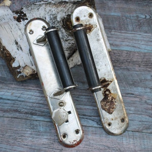 May include: Two silver door handles with black knobs. The handles have a decorative design and are mounted on a wooden surface.