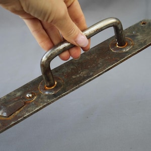 May include: A vintage, weathered metal door handle with a rectangular backplate and a curved handle. The handle and backplate show signs of rust and wear, suggesting age and use. The handle is held by a hand.