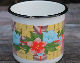 May include: A white enamel mug with a black rim and a floral pattern. The pattern features yellow, brown, red, blue, and green flowers on a yellow and brown plaid background.