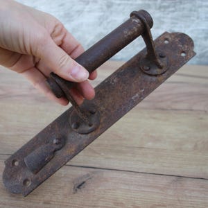 May include: An antique, rusty brown metal door handle with a long rectangular backplate and a cylindrical handle. The handle has a keyhole and decorative rivets. The metal shows signs of age and wear.