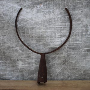 May include: A rusty metal tool with a curved top and a triangular handle. The tool is made of iron and has a weathered appearance. The background is a textured gray wall and a wooden surface.