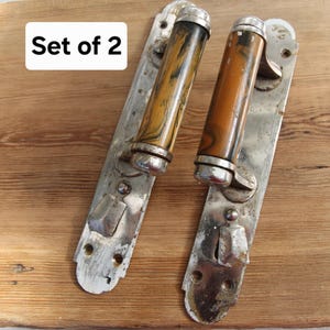 May include: A set of 2 door handles with rectangular silver-toned metal backplates and cylindrical brown and black marbled handles. The metal plates feature screw holes and a small latch mechanism. The text Set of 2 is visible on the image.