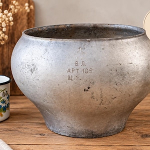 May include: A large, round, weathered cast aluminum pot with a wide opening and a narrow base. The exterior surface is textured and gray, featuring stamped Cyrillic text that reads 8 L. APT 105. A circular graphic in the corner indicates 8 Liters.