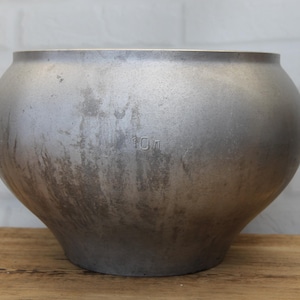 May include: A large, silver-colored metal cooking pot with a rounded body and a wide opening. The pot has a slightly distressed finish and the number "10l" is stamped on the side. It sits on a wooden surface.