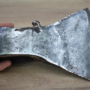 Peut inclure: Une tête de hache vintage avec une finition métallique patinée. La tête de hache est de couleur gris foncé avec des signes d'usure visibles, suggérant l'âge et l'utilisation. La conception présente une lame tranchante et angulaire et une base rectangulaire robuste.