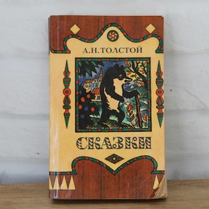 Puede incluir: Un libro de tapa dura con una cubierta de color madera beige y marrón que presenta una ilustración central de un oso negro en un paisaje. El texto A.H.TOЛCTOЙ y CKAЗKИ está impreso en negro. El borde incluye patrones geométricos verdes y rojos.