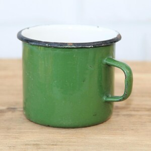 Puede incluir: Una taza de esmalte verde con interior blanco y borde negro. La taza tiene forma redondeada con asa lateral. La taza está sobre una superficie de madera.