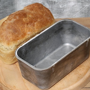 May include: A loaf of freshly baked bread sitting next to a silver loaf pan on a wooden cutting board.