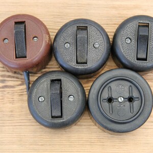 Può includere: Una collezione di interruttori della luce e una presa elettrica vintage. Gli interruttori rotondi con interruttore a levetta sono marroni e neri. La presa è nera con due fori e una vite centrale.