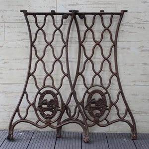 May include: Pair of antique cast iron sewing machine legs. The legs are a dark brown color with an ornate, openwork design. Each leg features an oval emblem with an eagle. The legs are sturdy and suitable for repurposing.