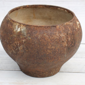May include: A rusty brown cast iron pot with a wide, round opening. The pot has a rough, textured surface and appears to be old and well-used.