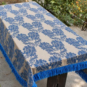 Op de afbeelding: Een rechthoekig tafelkleed met een beige basis en een herhalend blauw damast bloemenpatroon. De randen van de stof zijn afgewerkt met een dichte, bijpassende blauwe franje die langs de zijkanten van de tafel naar beneden hangt.